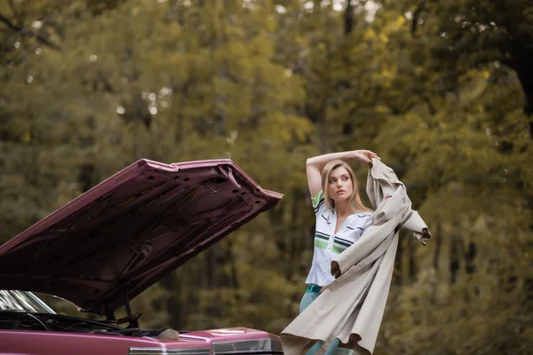 Молодая женщина надевает плащ, стоя рядом со сломанной винтажной машиной с открытым капотом — стоковое фото