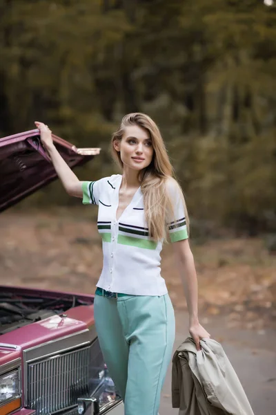 Young woman looking away while opening broken vintage car on blurred background — Stock Photo