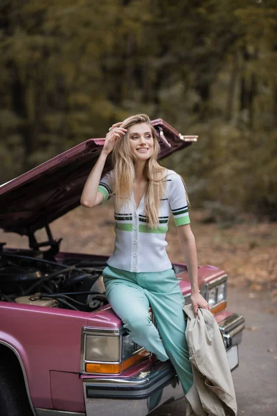 Femme souriante toucher les cheveux et regarder loin tout en étant assis sur une voiture cassée avec capuche ouverte — Photo de stock