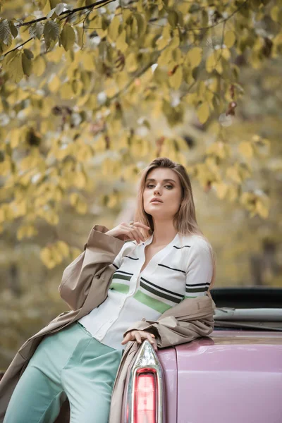 Junge stylische Frau in Umhang blickt in die Kamera, während sie in der Nähe von Cabriolet im Wald posiert — Stockfoto