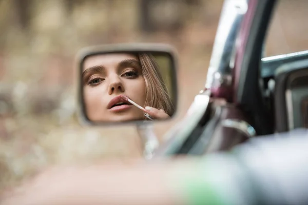 Mujer aplicando brillo de labios mientras mira el espejo de vista lateral en primer plano borroso - foto de stock