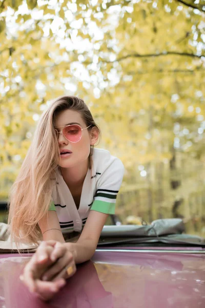 Young stylish woman in sunglasses leaning on hood of cabriolet on blurred foreground — Stock Photo