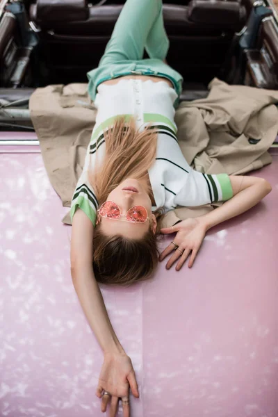 Overhead view of stylish woman in sunglasses relaxing while lying on car on blurred background — Stock Photo