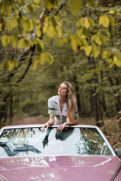 Giovane donna in occhiali da sole guardando altrove mentre si appoggia sul parabrezza dell'auto convertibile — Foto stock