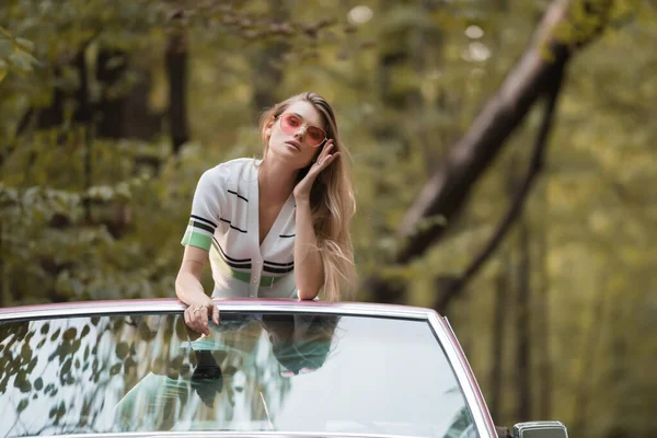 Jeune femme touchant des lunettes de soleil tout en s'appuyant sur le pare-brise de voiture convertible — Photo de stock