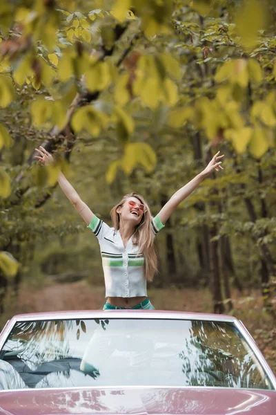 Mulher animado em óculos de sol de pé com as mãos no ar em cabriolet — Fotografia de Stock