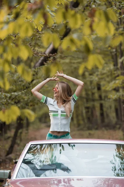 Mujer excitada en gafas de sol mirando hacia otro lado mientras está de pie con las manos por encima de la cabeza en cabriolet - foto de stock