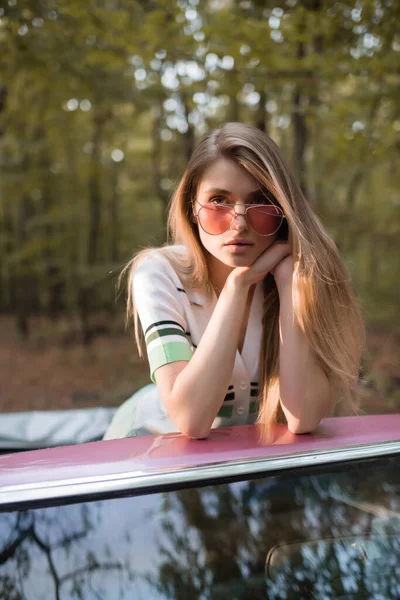 Stylische Frau mit Sonnenbrille, die die Hände vor dem Gesicht hält, während sie sich an die Windschutzscheibe des Cabrios lehnt — Stockfoto