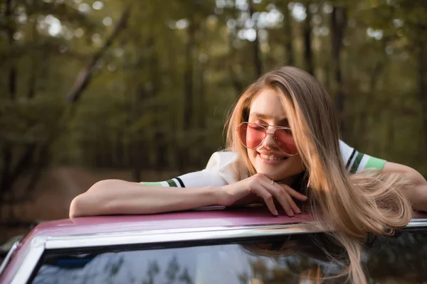 Mujer joven en gafas de sol sonriendo con los ojos cerrados mientras se apoya en el parabrisas de cabriolet - foto de stock