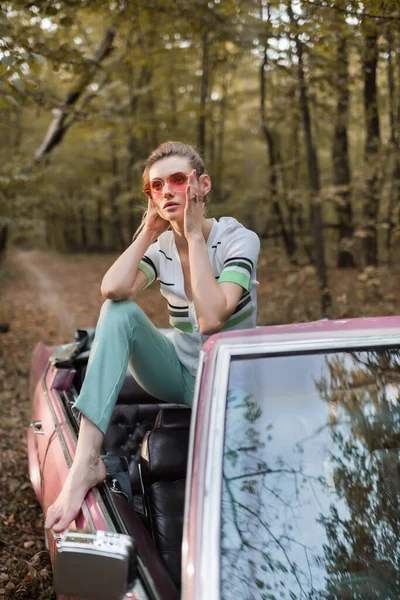 Стильна босоніжка торкається сонцезахисних окулярів під час позування в кабріолеті в лісі — стокове фото
