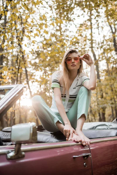 Stylish barefoot woman touching sunglasses while posing in cabriolet in forest on blurred foreground — Stock Photo