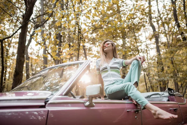Souriant pieds nus femme en lunettes de soleil posant en voiture convertible en forêt — Photo de stock