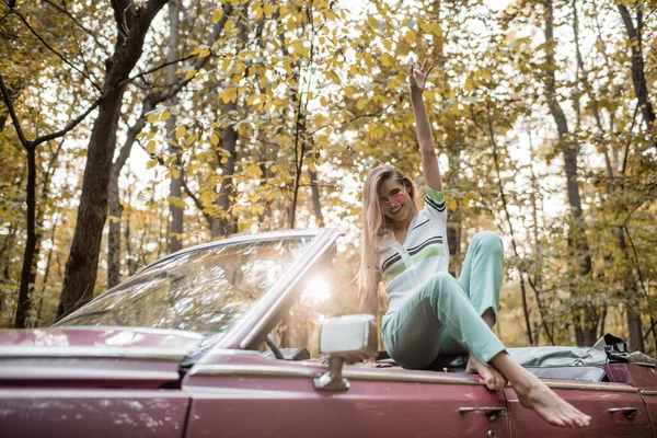 Joyful barefoot woman looking at camera while posing with hand in air in cabriolet — Stock Photo