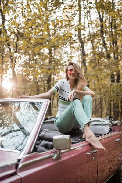 Fröhliche barfüßige Frau mit Brille blickt in die Kamera, während sie im Cabrio im Wald posiert — Stockfoto