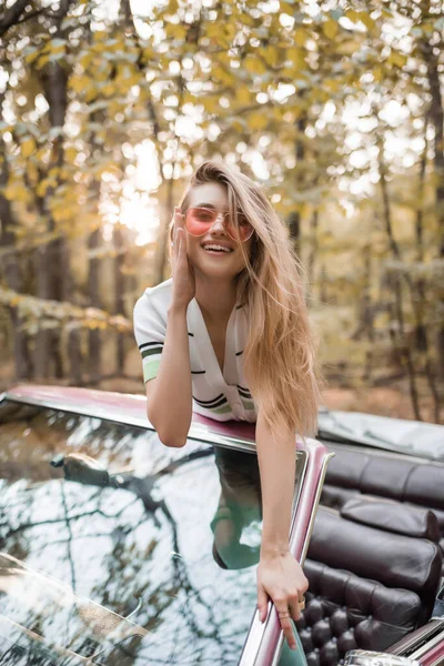 Sonriente mujer tocando gafas de sol mientras se apoya en el parabrisas de cabriolet y mirando a la cámara - foto de stock