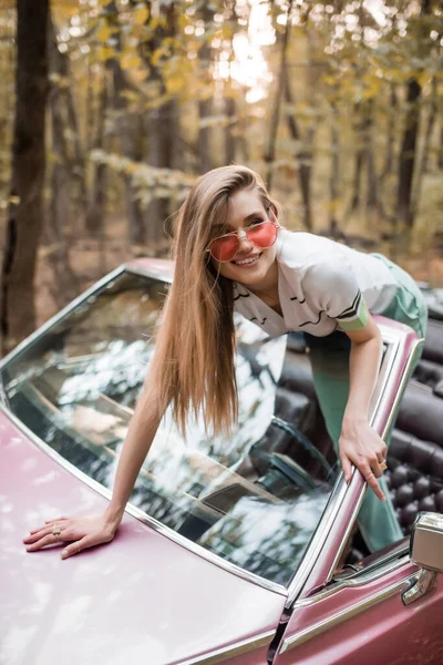 Glückliche Frau mit Sonnenbrille, die wegschaut, während sie sich an die Windschutzscheibe des Cabrios lehnt — Stockfoto