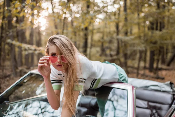 Stylische junge Frau berührt Sonnenbrille, während sie sich an Windschutzscheibe des Cabrios lehnt — Stockfoto