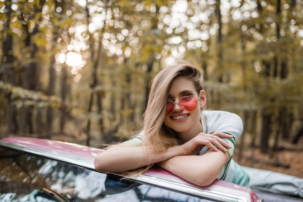 Gioiosa donna in occhiali da sole guardando la fotocamera mentre si appoggia sul parabrezza di cabriolet con braccia incrociate — Foto stock