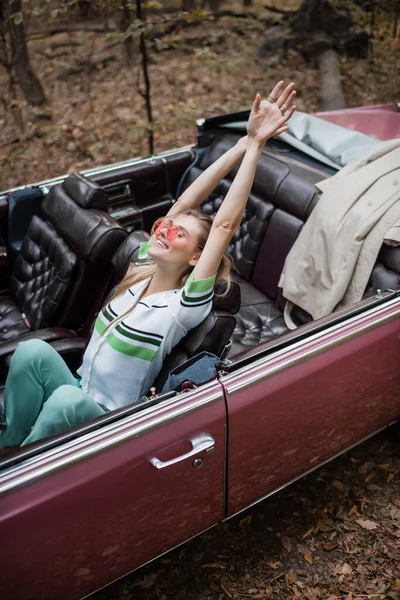 Vista de ángulo alto de la mujer alegre relajándose mientras está sentado en cabriolet retro y estirando las manos - foto de stock