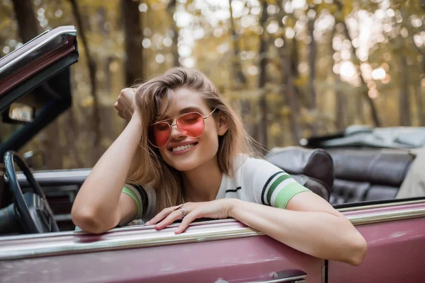 Happy woman in sunglasses holding hand near head while sitting in cabriolet with closed eyes — Stock Photo