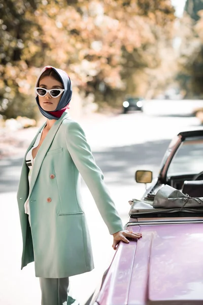Mujer de moda en pañuelo y gafas de sol de pie en la carretera cerca de cabriolet - foto de stock