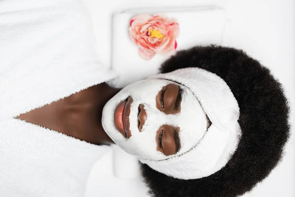 Вид сверху на кудрявую африканскую женщину, лежащую с маской для лица рядом с цветочком в спа-салоне — стоковое фото
