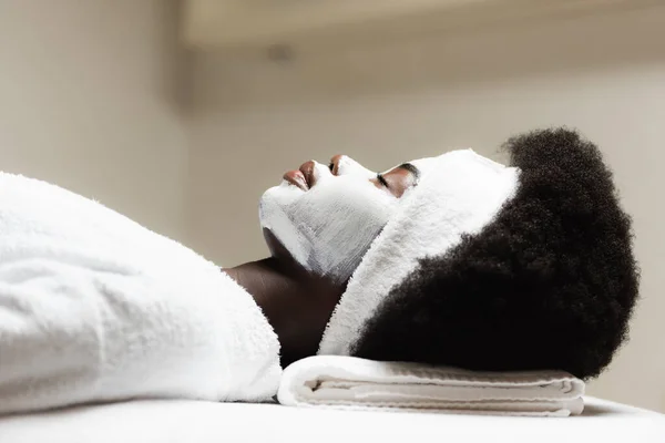 Vue latérale de la femme afro-américaine bouclée couchée avec masque facial sur fond flou — Photo de stock