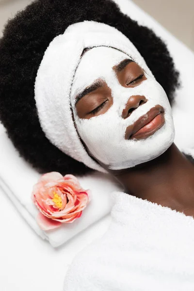 Retrato de mulher afro-americana encaracolada com cabeça branca e máscara facial deitada perto da flor — Fotografia de Stock