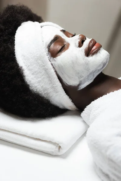 Nahaufnahme der lockigen Afroamerikanerin mit weißem Stirnband und Gesichtsmaske, die im Wellness-Salon liegt — Stockfoto
