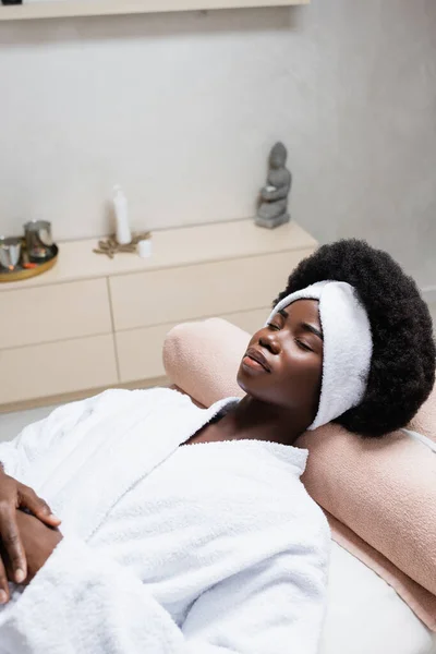 Mujer afroamericana en bata de baño descansando, mientras que la colocación en la mesa de masaje en el salón de spa sobre fondo borroso - foto de stock