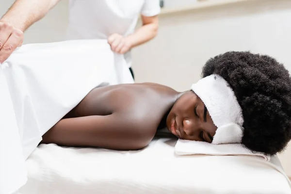 Masseur couvrant femme afro-américaine avec drap de lit dans le salon de spa, bannière — Photo de stock