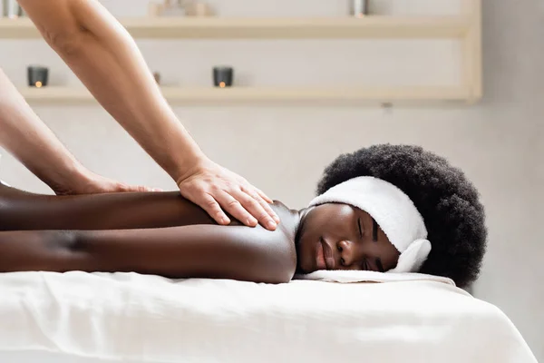 Massaggiatore massaggio africano americano donna indossare bianco fascia in spa salone su sfondo sfocato — Foto stock