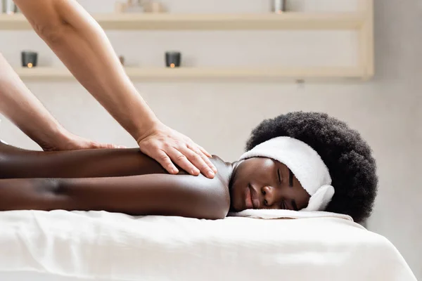 Masajista masajeando mujer afroamericana con diadema blanca en salón de spa sobre fondo borroso - foto de stock