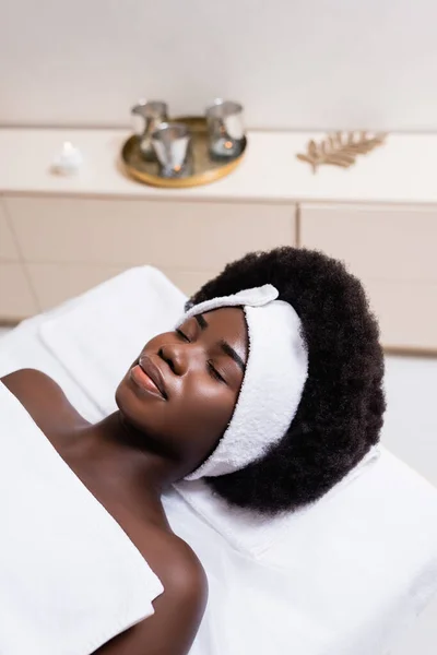 Afroamerikanerin in weißem Haarband mit Bettlaken bedeckt auf Massagetisch im Wellness-Salon auf verschwommenem Hintergrund liegend — Stockfoto