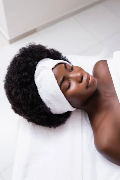 Overhead view of african american woman in white headband lying on massage table in spa salon — Stock Photo