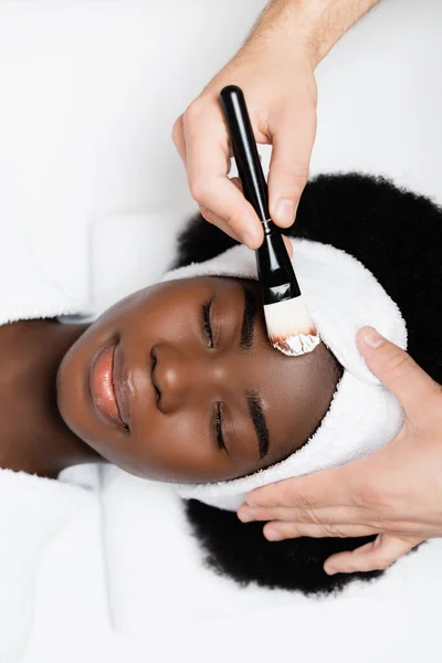 Vue du dessus de la femme afro-américaine souriante en bandeau blanc couché près de l'homme avec une brosse cosmétique dans un salon de spa — Photo de stock