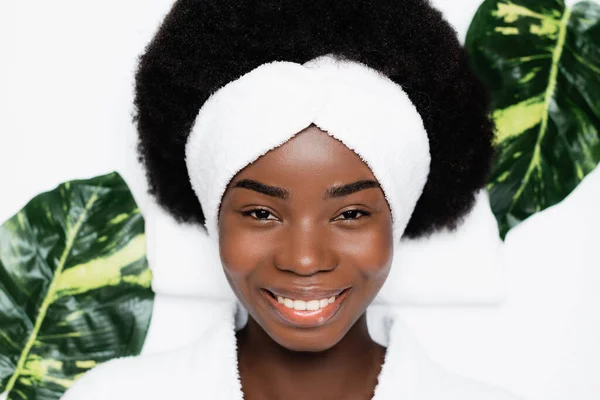 Vista dall'alto della donna afro-americana sorridente con fascia bianca che guarda la fotocamera, mentre riposa vicino alle foglie verdi nel salone spa — Foto stock