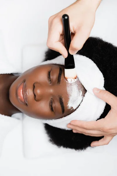 Wellness-Therapeutin trägt Gesichtsmaske auf der Stirn einer Afroamerikanerin mit Kosmetikbürste im Wellness-Salon auf — Stockfoto