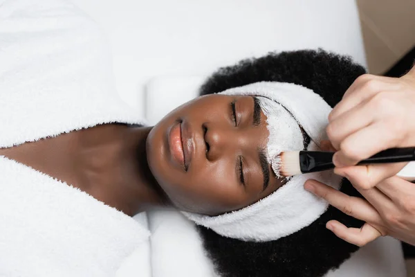 Terapeuta de spa que aplica mascarilla facial con cepillo cosmético en la frente de una mujer afroamericana que usa abrigo de rizo en el salón de spa - foto de stock