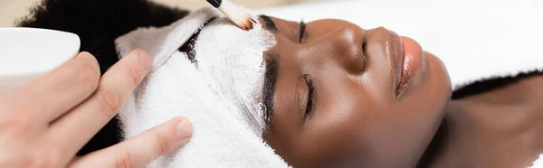 Close up view of spa therapist applying face mask with cosmetic brush on forehead of african american woman in white headband, banner — Stock Photo