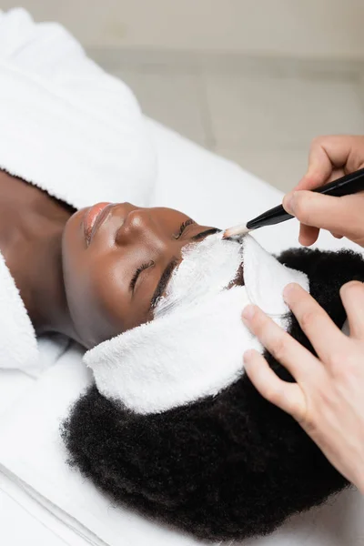 Overhead view of spa therapist applying face mask with cosmetic brush on forehead of african american woman in spa salon — Stock Photo