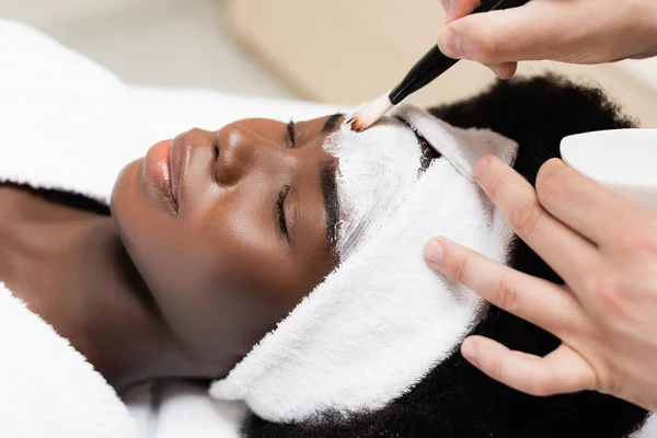 Vista da vicino dell'uomo che applica maschera viso con pennello cosmetico sulla fronte della donna afro-americana nel salone spa — Foto stock