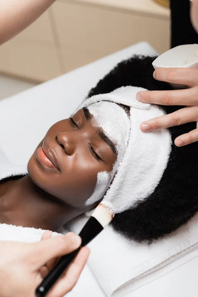 Vista de perto do terapeuta spa aplicando máscara facial com escova cosmética na bochecha da mulher afro-americana deitada no salão de spa — Fotografia de Stock