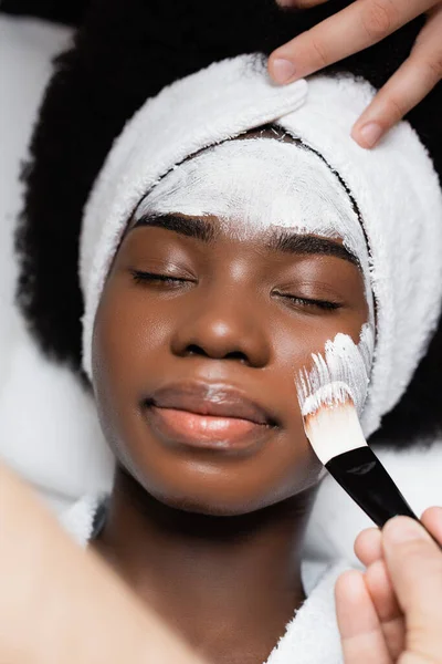 Vista dall'alto della donna afro-americana sdraiata, mentre il terapeuta spa applica la maschera facciale sulla guancia con pennello cosmetico nel salone spa — Foto stock