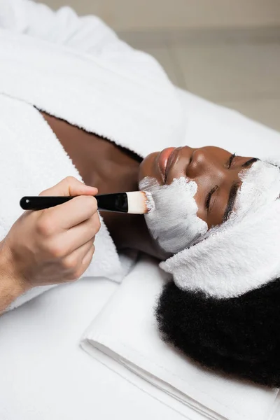 Mulher afro-americana positiva na cabeça deitada perto de terapeuta spa aplicando máscara facial na bochecha no salão de spa — Fotografia de Stock