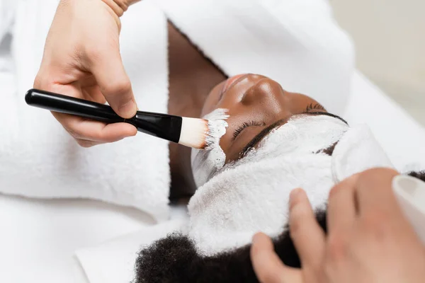 Afroamerikanerin mit Stirnband liegt in der Nähe von Wellness-Therapeutin und setzt Gesichtsmaske auf Wange im Wellness-Salon auf — Stockfoto