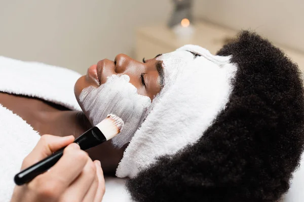 Nahaufnahme einer Afroamerikanerin mit Stirnband, die in der Nähe eines Mannes liegt, der im Wellness-Salon auf verschwommenem Hintergrund eine Gesichtsmaske auf die Wange aufträgt — Stockfoto