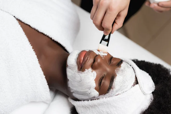 Close up vista da mulher afro-americana deitado perto de terapeuta spa aplicando creme facial na bochecha no salão de spa — Fotografia de Stock