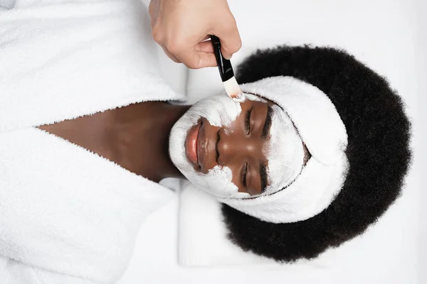 Top view of african american woman with closed eyes, wearing bathrobe, while lying near man applying face mask on cheek in spa salon — Stock Photo
