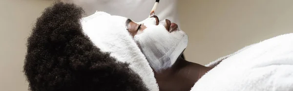 Side view of african american woman lying near man applying face mask with cosmetic brush on nose in spa salon, banner — Stock Photo
